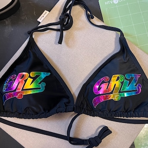 Peut inclure: Un haut de bikini noir avec un lien &agrave; nouer autour du cou et des bonnets triangulaires. Chaque bonnet affiche le texte "GRIZ" dans une finition holographique et paillet&eacute;e aux couleurs vives, variant entre le violet, bleu, vert, jaune et rouge.