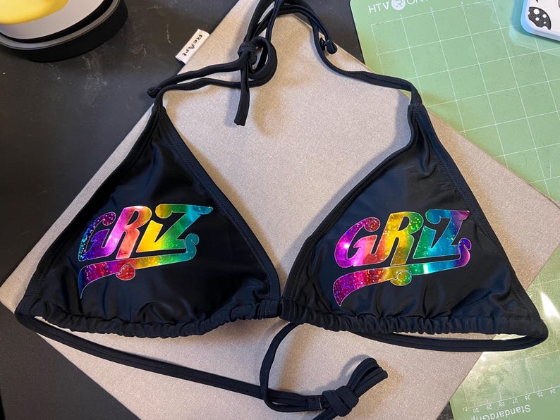 Peut inclure: Un haut de bikini noir avec un lien &agrave; nouer autour du cou et des bonnets triangulaires. Chaque bonnet affiche le texte "GRIZ" dans une finition holographique et paillet&eacute;e aux couleurs vives, variant entre le violet, bleu, vert, jaune et rouge.
