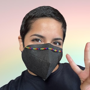 May include: A person wearing a black face mask with a rainbow beaded chain. The mask is covering the person's nose and mouth. The person is looking at the camera and has a peace sign up.