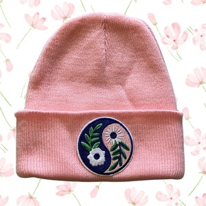 May include: Pink knitted beanie with a yin yang symbol embroidered on the front. The symbol features a white flower and green leaves on a blue background.