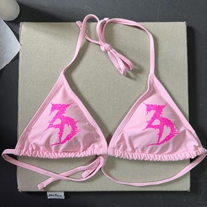 Peut inclure: Un haut de bikini rose clair avec une silhouette triangulaire et de fines bretelles r&eacute;glables &agrave; nouer au cou et dans le dos. L'avant de chaque bonnet pr&eacute;sente un motif graphique &agrave; paillettes rose vif avec des lettres stylis&eacute;es en forme de 35.