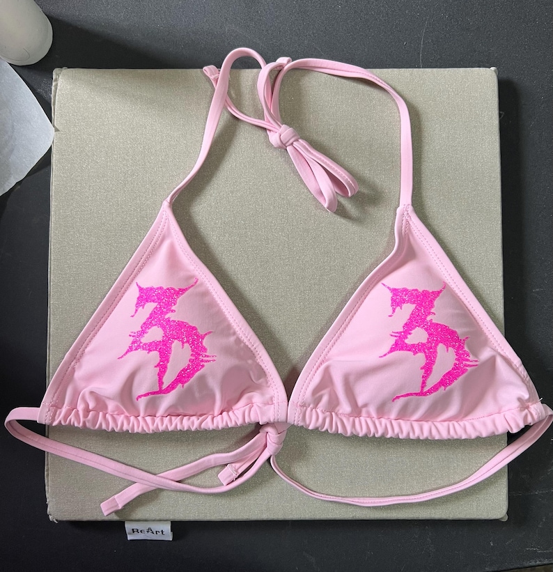 Peut inclure: Un haut de bikini rose clair avec une silhouette triangulaire et de fines bretelles r&eacute;glables &agrave; nouer au cou et dans le dos. L'avant de chaque bonnet pr&eacute;sente un motif graphique &agrave; paillettes rose vif avec des lettres stylis&eacute;es en forme de 35.