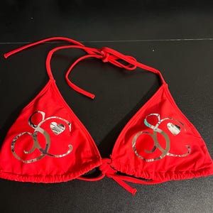 Peut inclure: Un haut de bikini rouge vif compos&eacute; de deux bonnets triangulaires avec des liens &agrave; nouer r&eacute;glables au cou et dans le dos. Chaque bonnet pr&eacute;sente un grand logo m&eacute;tallique argent&eacute; r&eacute;fl&eacute;chissant form&eacute; de lettres cursives et d'un petit c&oelig;ur.