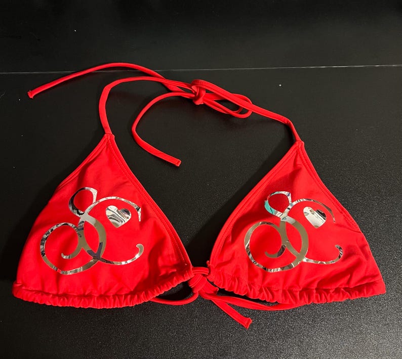 Peut inclure: Un haut de bikini rouge vif compos&eacute; de deux bonnets triangulaires avec des liens &agrave; nouer r&eacute;glables au cou et dans le dos. Chaque bonnet pr&eacute;sente un grand logo m&eacute;tallique argent&eacute; r&eacute;fl&eacute;chissant form&eacute; de lettres cursives et d'un petit c&oelig;ur.