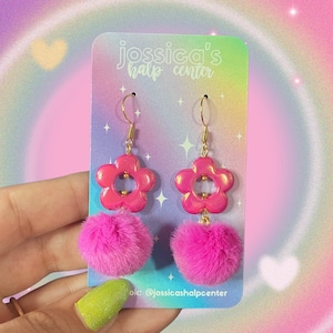 May include: Pink flower-shaped earrings with a gold hook and a pink fluffy pom-pom charm. The earrings are on a white card with the text "jossica's half center" and "ok: @jossicashalpcenter".