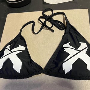 Peut inclure: Un haut de bikini noir orn&eacute; d'un graphisme blanc en forme de lettre X sur chaque bonnet triangulaire. Le v&ecirc;tement comprend de fines attaches en tissu noir au niveau du cou et du dos, avec une silhouette triangulaire classique et un tissu noir uni.