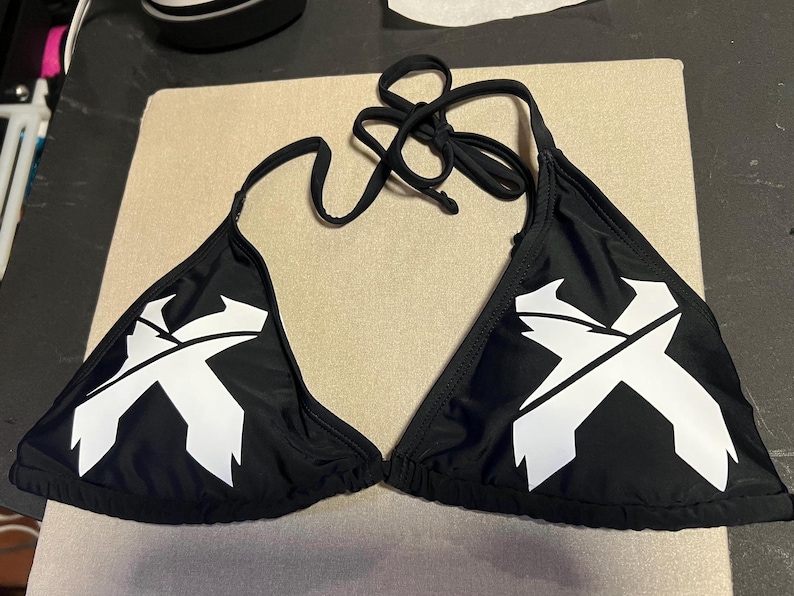 Peut inclure: Un haut de bikini noir orn&eacute; d'un graphisme blanc en forme de lettre X sur chaque bonnet triangulaire. Le v&ecirc;tement comprend de fines attaches en tissu noir au niveau du cou et du dos, avec une silhouette triangulaire classique et un tissu noir uni.