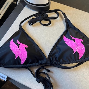 Peut inclure: Un haut de bikini noir orn&eacute; d'un motif de ph&eacute;nix rose vif &agrave; paillettes sur chaque bonnet triangulaire. Le v&ecirc;tement comprend de fines bretelles noires &agrave; nouer au cou et dans le dos, offrant un ajustement r&eacute;glable avec une silhouette classique.
