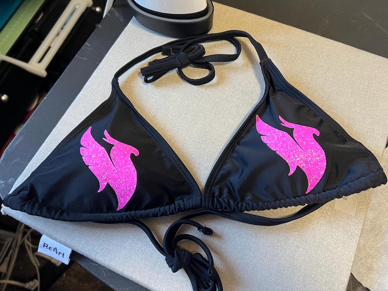 Peut inclure: Un haut de bikini noir orn&eacute; d'un motif de ph&eacute;nix rose vif &agrave; paillettes sur chaque bonnet triangulaire. Le v&ecirc;tement comprend de fines bretelles noires &agrave; nouer au cou et dans le dos, offrant un ajustement r&eacute;glable avec une silhouette classique.