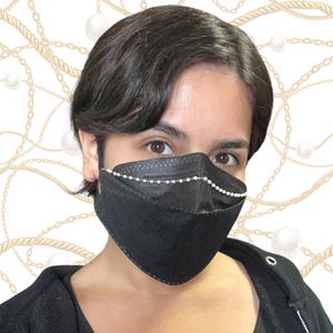 May include: A black face mask with a row of small, white pearls along the top edge. The mask is made of a textured material and is worn by a person. The background features a pattern of gold chains, ropes, and white pearls.