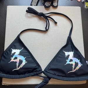 Peut inclure: Un haut de bikini noir compos&eacute; de deux bonnets triangulaires avec de fines attaches r&eacute;glables au cou et au dos. Chaque bonnet pr&eacute;sente un grand motif graphique iris&eacute; de style m&eacute;tallique avec un lettrage stylis&eacute; aux bords dentel&eacute;s.