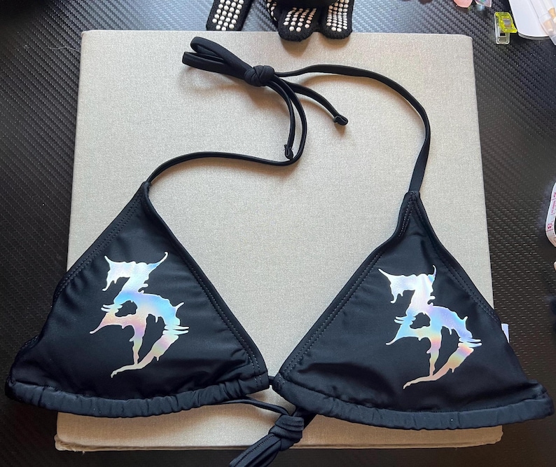 Peut inclure: Un haut de bikini noir compos&eacute; de deux bonnets triangulaires avec de fines attaches r&eacute;glables au cou et au dos. Chaque bonnet pr&eacute;sente un grand motif graphique iris&eacute; de style m&eacute;tallique avec un lettrage stylis&eacute; aux bords dentel&eacute;s.