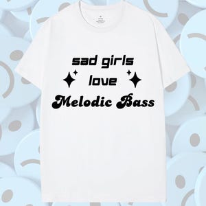 May include: White t-shirt featuring the text "sad girls love Melodic Bass" in black. The text is centered, with two black diamond shapes. The shirt has a crew neck and short sleeves.