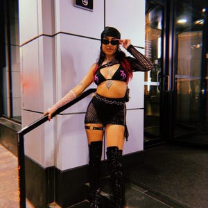 Peut inclure: Un haut asym&eacute;trique noir en r&eacute;sille et une mini-jupe en r&eacute;sille assortie avec une ceinture &agrave; motifs d'&eacute;toiles. La tenue comprend un haut de bikini triangle noir avec des d&eacute;tails graphiques roses, un foulard noir, des lunettes et des bottes.