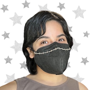 May include: A person wearing a black face mask with a silver chain featuring star-shaped charms. The chain is attached to the mask and goes over the ears.