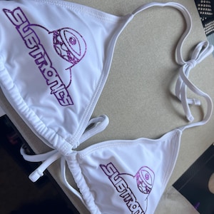 Peut inclure: Un haut de bikini blanc orn&eacute; d'un texte violet &agrave; paillettes indiquant Subtronics, accompagn&eacute; d'un motif graphique violet &agrave; paillettes repr&eacute;sentant un &oelig;il de cyclope. Le v&ecirc;tement poss&egrave;de des liens ajustables au cou et au dos.