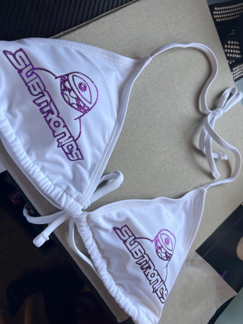 Peut inclure: Un haut de bikini blanc orn&eacute; d'un texte violet &agrave; paillettes indiquant Subtronics, accompagn&eacute; d'un motif graphique violet &agrave; paillettes repr&eacute;sentant un &oelig;il de cyclope. Le v&ecirc;tement poss&egrave;de des liens ajustables au cou et au dos.