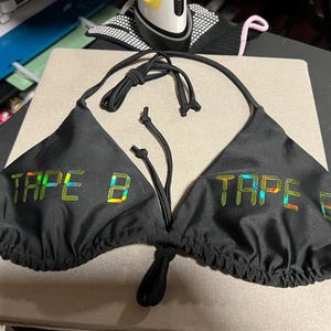 Peut inclure: Un haut de bikini noir compos&eacute; de deux bonnets triangulaires avec de fines bretelles &agrave; nouer. Le texte TAPE B est imprim&eacute; sur chaque bonnet dans une finition holographique r&eacute;fl&eacute;chissante, iris&eacute;e et multicolore sur le tissu noir uni.