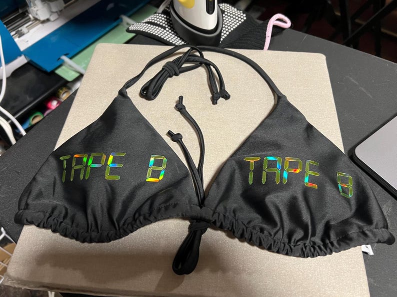 Peut inclure: Un haut de bikini noir compos&eacute; de deux bonnets triangulaires avec de fines bretelles &agrave; nouer. Le texte TAPE B est imprim&eacute; sur chaque bonnet dans une finition holographique r&eacute;fl&eacute;chissante, iris&eacute;e et multicolore sur le tissu noir uni.