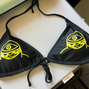 Peut inclure: Un haut de bikini noir orn&eacute; d'un imprim&eacute; graphique jaune vif repr&eacute;sentant un &oelig;il stylis&eacute; sur chaque bonnet triangulaire. Le v&ecirc;tement comprend de fines bretelles noires &agrave; nouer autour du cou et un lien assorti au centre, en tissu lisse.