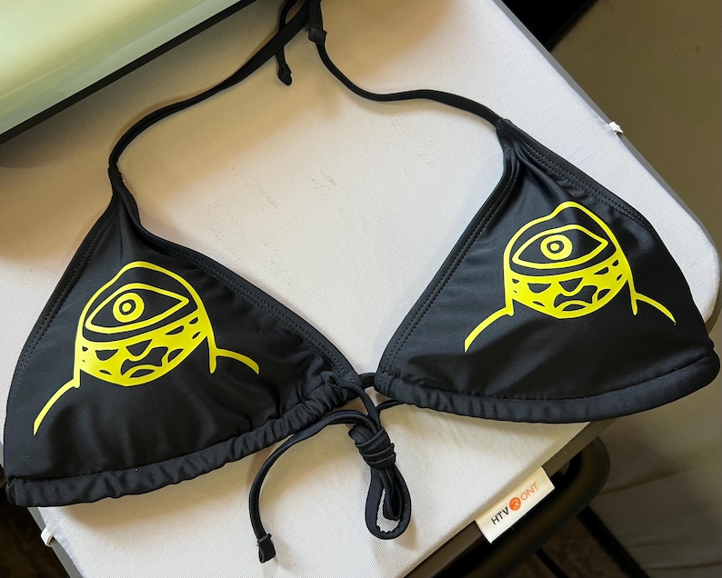Peut inclure: Un haut de bikini noir orn&eacute; d'un imprim&eacute; graphique jaune vif repr&eacute;sentant un &oelig;il stylis&eacute; sur chaque bonnet triangulaire. Le v&ecirc;tement comprend de fines bretelles noires &agrave; nouer autour du cou et un lien assorti au centre, en tissu lisse.
