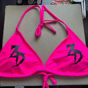 Peut inclure: Un haut de bikini rose n&eacute;on dot&eacute; d'un design tour de cou avec de fines bretelles r&eacute;glables et une fermeture &agrave; nouer dans le dos. L'avant de chaque bonnet triangulaire est imprim&eacute; d'un logo graphique noir, dentel&eacute; et stylis&eacute; en forme de ZD.