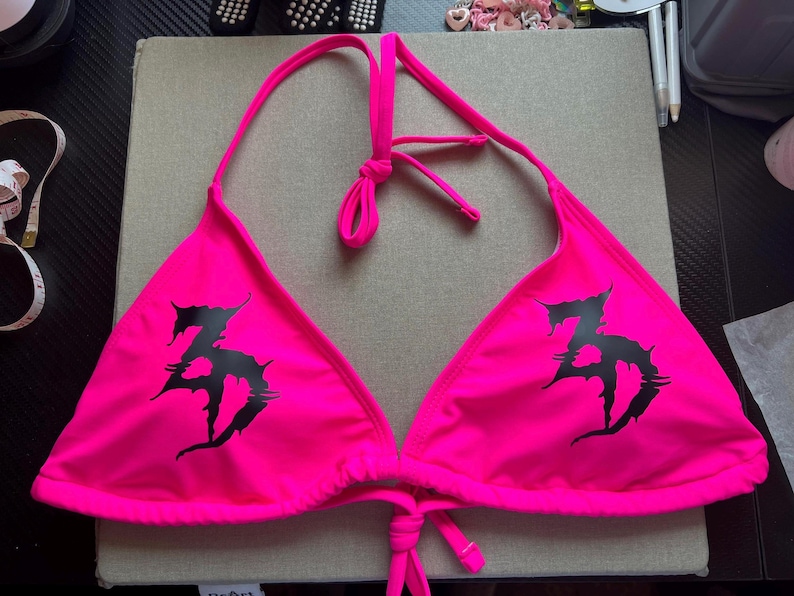 Peut inclure: Un haut de bikini rose n&eacute;on dot&eacute; d'un design tour de cou avec de fines bretelles r&eacute;glables et une fermeture &agrave; nouer dans le dos. L'avant de chaque bonnet triangulaire est imprim&eacute; d'un logo graphique noir, dentel&eacute; et stylis&eacute; en forme de ZD.