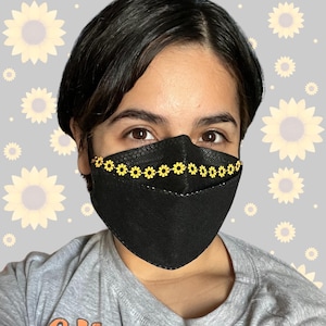 May include: Black face mask with a decorative row of yellow sunflowers. The mask is made of a textured material and has a stitched design. The background features a pattern of sunflowers.