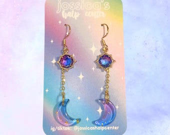 May include: A pair of gold dangle earrings with a blue and purple iridescent star charm and a crescent moon charm. The earrings are on a card that says "jossica's half center" and "ig/tiktok: @jossicashalpcente"