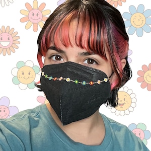 May include: A black face mask with a chain of small, colourful daisy flower beads. The person wearing the mask has dark hair with pink highlights. The background features smiling flower graphics.