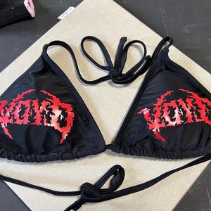 Peut inclure: Un haut de bikini noir compos&eacute; de deux bonnets triangulaires avec de fines bretelles r&eacute;glables. L'avant de chaque bonnet pr&eacute;sente le mot VOID imprim&eacute; dans une police rouge dentel&eacute;e et stylis&eacute;e. Le v&ecirc;tement est en tissu noir uni.