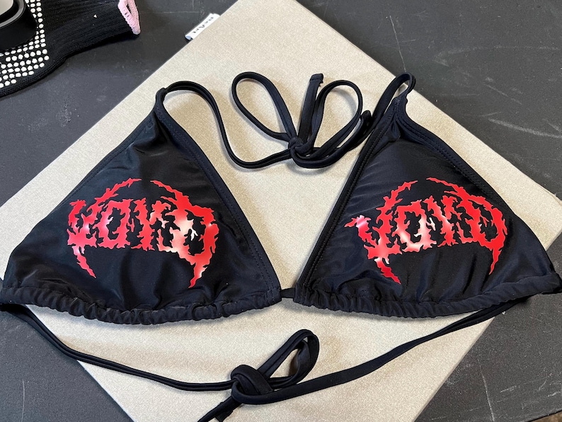 Peut inclure: Un haut de bikini noir compos&eacute; de deux bonnets triangulaires avec de fines bretelles r&eacute;glables. L'avant de chaque bonnet pr&eacute;sente le mot VOID imprim&eacute; dans une police rouge dentel&eacute;e et stylis&eacute;e. Le v&ecirc;tement est en tissu noir uni.