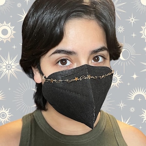 May include: A black face mask with a gold chain adorned with stars and crescent moons. The chain is attached to the mask's ear loops.