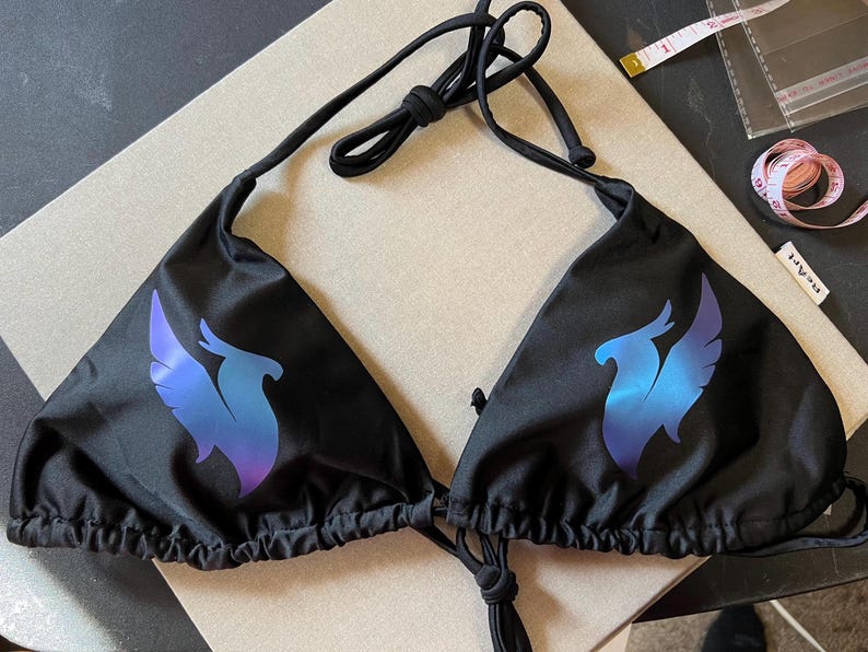Peut inclure: Un haut de bikini noir orn&eacute; d'un motif de ph&eacute;nix en d&eacute;grad&eacute; bleu et violet sur chaque bonnet triangulaire. Le v&ecirc;tement comporte de fines bretelles noires &agrave; nouer au cou et dans le dos. Un ruban &agrave; mesurer blanc indique 5 cm.