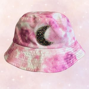 May include: Pink tie-dye bucket hat with a crescent moon design embellished with iridescent rhinestones.