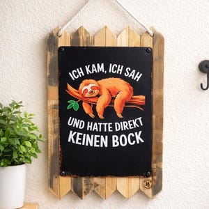 May include: A rectangular wall sign featuring a black metal plate mounted on a vertical wooden picket frame. The plate displays an illustration of an orange sloth sleeping on a branch with the German text: ICH KAM, ICH SAH UND HATTE DIREKT KEINEN BOCK.