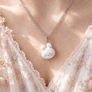 Könnte beinhalten: Eine silberne Halskette mit einem Anhänger in Form eines weißen Hasen mit rosa Ohren und Wangen. Der Hasenanhänger hat ein kleines, einfaches Gesicht. Die Halskette wird vor einem floral gemusterten Hintergrund präsentiert.