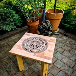 Könnte beinhalten: Ein quadratischer Beistelltisch aus Holz mit einer hellen Naturholzoberfläche. Die Tischplatte zeigt ein kreisförmiges, dunkel lasergraviertes Design einer Person mit Federschmuck, umgeben von komplizierten geometrischen Mustern.