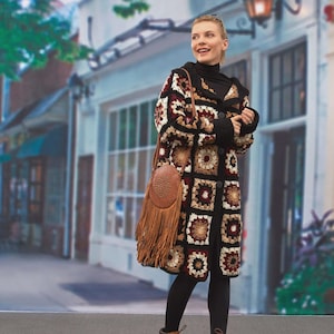May include: A long, crocheted coat in a patchwork design of black, cream, and burgundy squares. The coat has a hood and is buttoned up. A brown leather shoulder bag with fringe hangs from the shoulder.