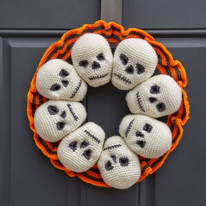 May include: A Halloween wreath made of crocheted white skulls with black eyes and mouths. The skulls are arranged in a circle around a base of orange and black striped fabric.