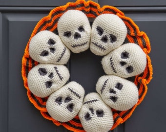 May include: A Halloween wreath made of crocheted white skulls with black eyes and mouths. The skulls are arranged in a circle around a base of orange and black striped fabric.