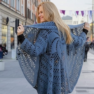Peut inclure: Un cardigan en crochet bleu avec un motif en dentelle. Le cardigan a un col large et ouvert et des manches longues. Le cardigan est porté par une personne dans un environnement urbain.