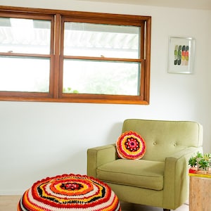 May include: A large, round, colorful crocheted floor cushion with red, orange, yellow, white, and black stripes. A smaller, round, crocheted pillow with a similar color scheme is on a green armchair in the background.