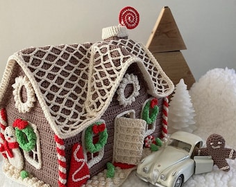 May include: A crocheted gingerbread house with red and white candy cane accents, green wreaths, and a red and white candy swirl on top. The house is sitting on a white surface with a toy car and gingerbread person.
