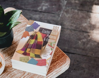 Puede incluir: Un libro titulado "101 Crochet Stitch Patterns & Edgings" con una manta de crochet colorida en la portada. El libro está abierto en una página que muestra diferentes patrones de puntadas de crochet.