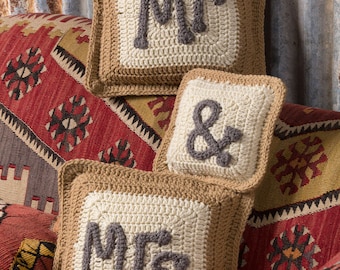 Op de afbeelding: Vierkante gehaakte kussens in beige en crème, met de woorden "MR", "MRS" en een ampersand in grijs. De kussens hebben een rand en staan tegen een achtergrond van een patroonkleed. Afmetingen in centimeters.