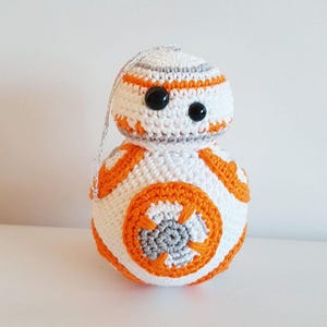 May include: A handmade crochet BB-8 droid ornament. The spherical figure is white with orange and gray accents, including a large orange circle on the lower half. It has black button eyes and a silver string for hanging. A Star Wars themed item.