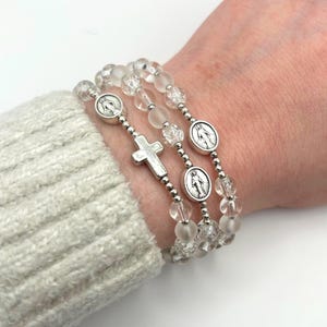 May include: Three clear beaded bracelets with silver accents. Each bracelet features a silver cross or a silver medallion with a religious figure. The bracelets are worn on a wrist, showcasing the delicate design and craftsmanship.