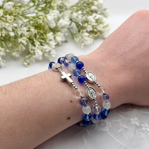 Könnte beinhalten: Drei Armbänder mit blauen, weißen und transparenten Perlen. Ein Armband hat ein silbernes Kreuz, die anderen beiden silberne Medaillons. Die Armbänder werden am Handgelenk getragen.