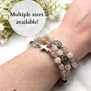 May include: A multi-strand beaded bracelet featuring translucent light-toned beads, small silver-colored spacers, a silver-colored cross charm, and two oval silver-colored religious medals. The image includes the text Multiple sizes available!